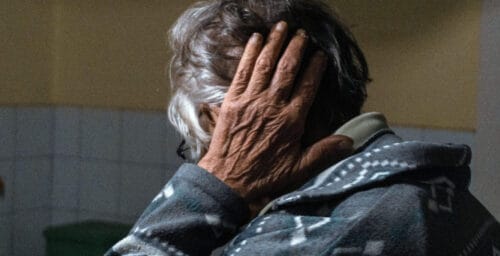 En äldre kvinna sitter med ryggen vänd emot kameran och håller sig för nacken